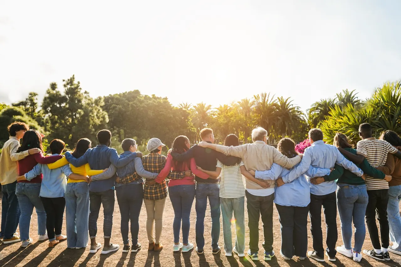 Um grupo diverso e multigeracional de pé, de braços dados, olhando juntos para o pôr do sol
